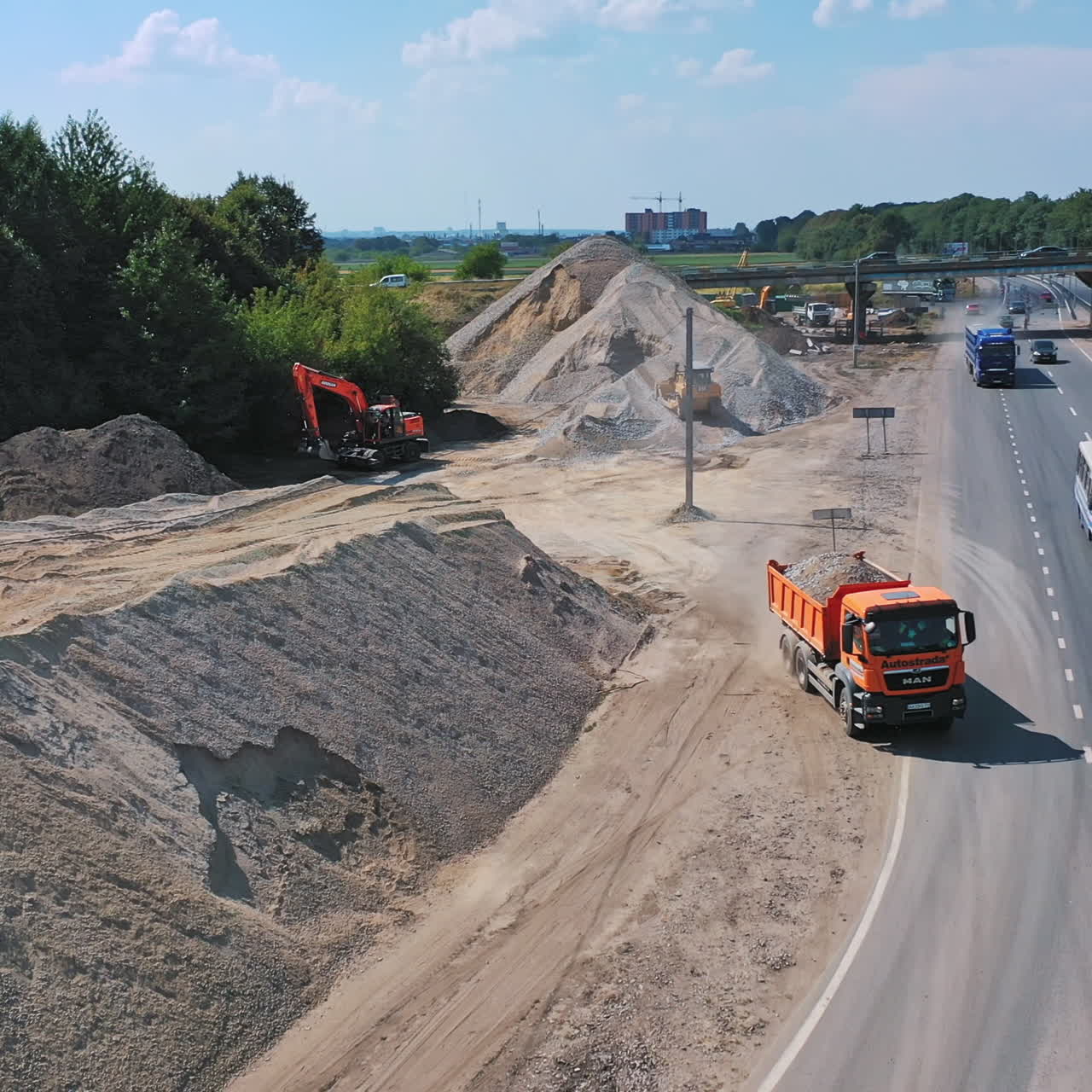 Pile of rubbles near the road. Truck full of stones moving out into the highway. Many cars driving on the road. Aerial view.