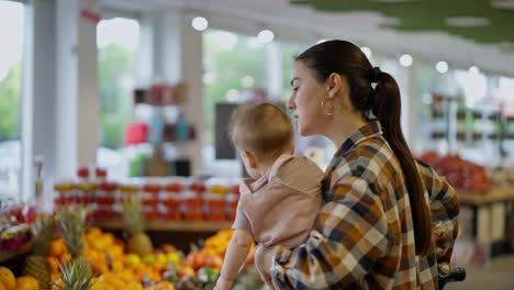 close-up a confident brunette girl in a plaid shirt together with her small child in her arms examines fruits in the supermarket department