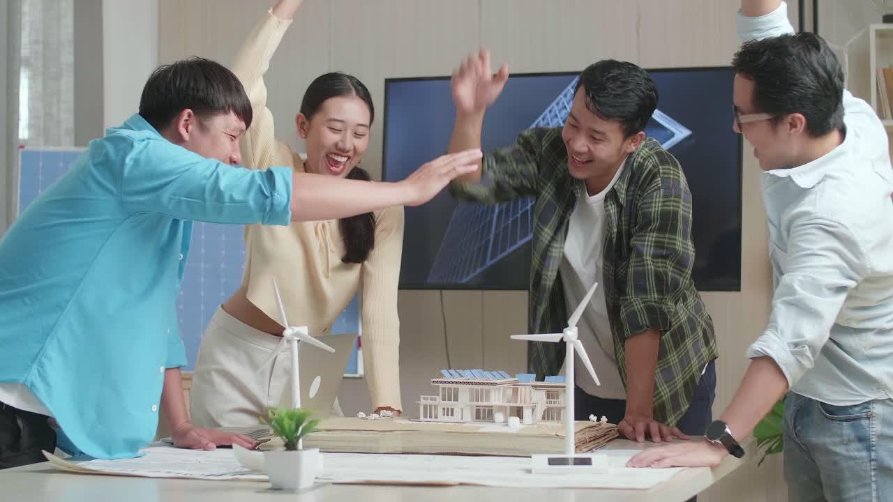 아시아 엔지니어 그룹이 사무실에서 태양 전지판을 가진 작은 집 모델을 작업하는 동안 함께 손을 고 있습니다.