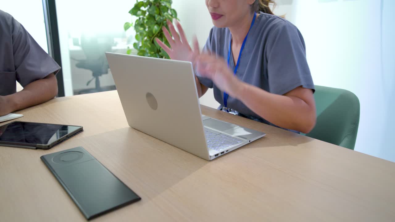 Vielfältiges Team in OP-Kleidung mit blauen Schlüsselbändern am Tisch, Frau bemerkt Laptop-Daten, präsentiert Plan