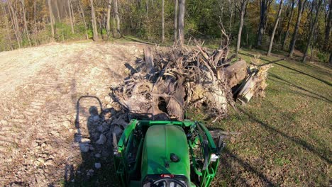 punto de vista de ángulo alto en un pequeño tractor verde usando horquillas elevadoras para recoger un tocón de árbol de un montón y mover escombros cerca de los bosques a principios de otoño