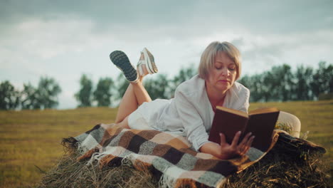 woman with leg raised on hay bale, checkered blanket beneath, reading book in an open field, strand of hay in mouth, sun hat beside her hand, enjoying peaceful moment in nature