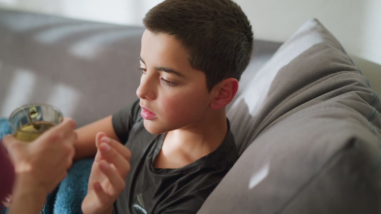 Close-up of sick boy lying on couch receiving tea from caregiver, he gently gets up to collect the cup, showing a moment of care and concern while recovering
