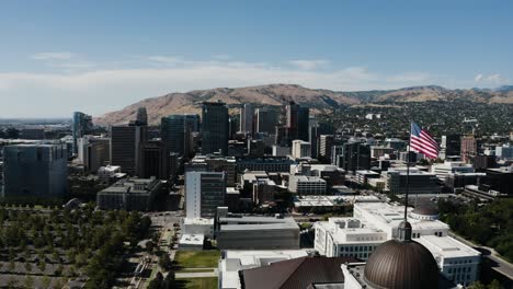 Toma-De-Drone-De-La-Bandera-Estadounidense-Ondeando-Sobre-Salt-Lake-City-En-Un-Día-Soleado