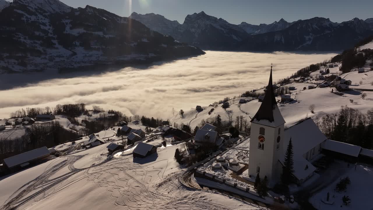 Aerial drone view of Amden village, highlighting its beautiful church with Lake Walensee in the background, partially covered by clouds.