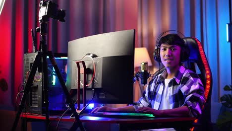 asian boy streamer smiling to camera while playing game over network on personal computer. live stream video game, desk illuminated by rgb led strip light