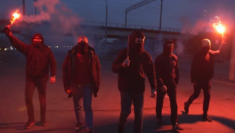 group of young men in balaclavas with red burning signal flare walking on the road under the bridge, slow motion