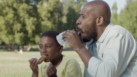 black man and boy sitting on bench in park, enjoying tasty snack