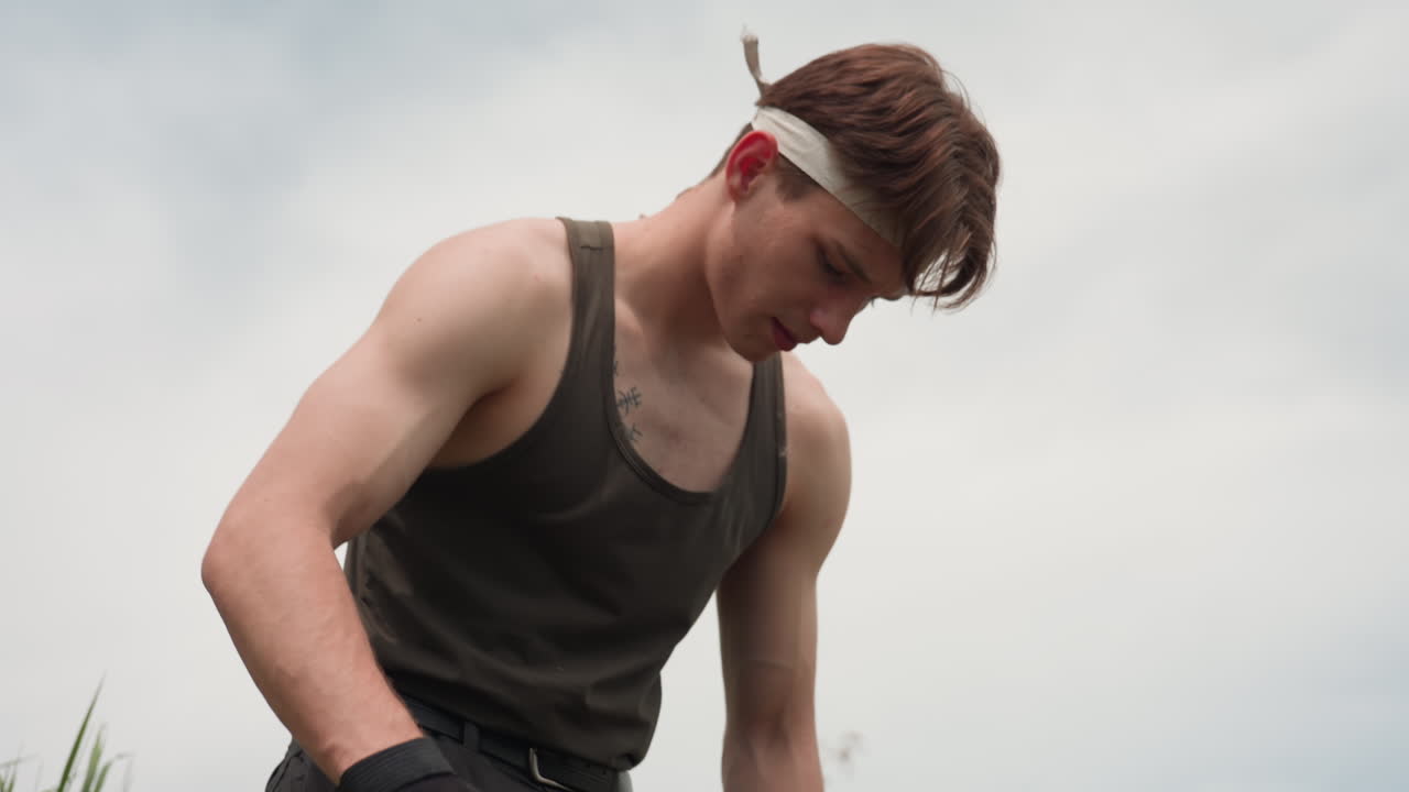 warrior in tank top and cargo pants brushing stubborn sand from pant leg under bright sky on grassy riverbank, tactical belt, gloved hand, loose soil, distant treeline and soft breeze