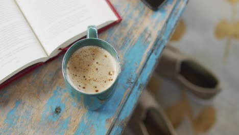 worn, painted wooden table top with notebook and cup of coffee with milk, slippers below on floor