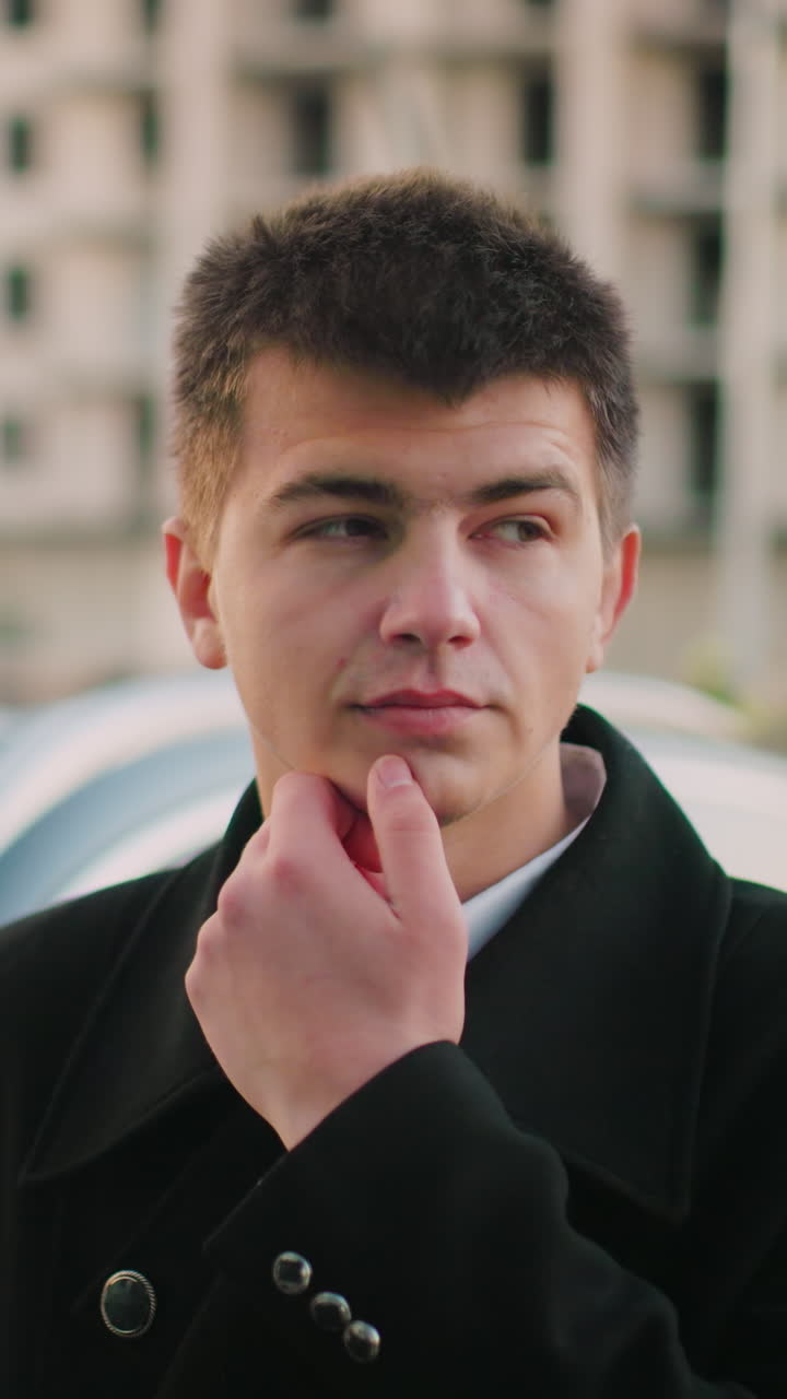 Business man standing close to his car, hand on jaw, thoughtfully looking ahead, with office building and parked cars in background