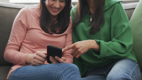 happy biracial mother and adult daughter using smartphone on couch at home, slow motion