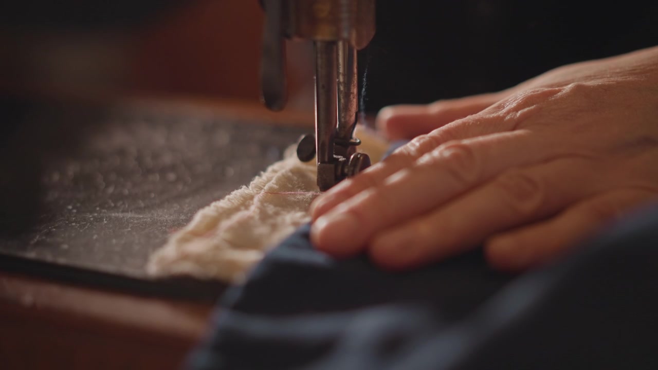 Cucito artigianale accurato sotto una luce calda. Un artigiano crea meticolosamente cuciture perfette su un bordo di lana blu navy. Artigiano esperto che si concentra intensamente sulla cucitura e la rifinitura di un capo in lana blu navy.
