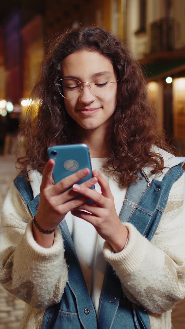 Ragazza adolescente felice e sorridente con gli occhiali che usa lo smartphone e scrive messaggi di testo in una strada urbana