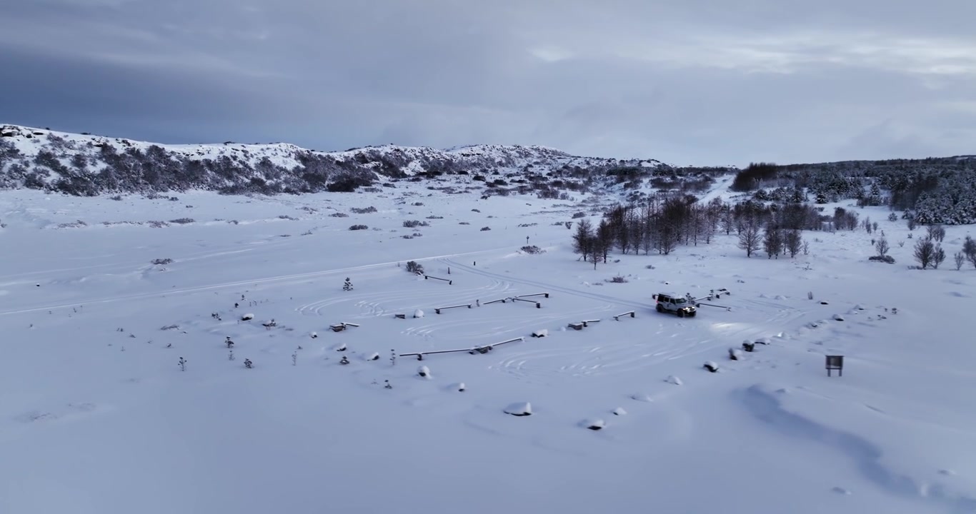 Wide drone view of a snowy Heiðmörk parking area in Iceland, with a 4x4 near parking barriers, frozen lake, winter hills, and cloudy sky.