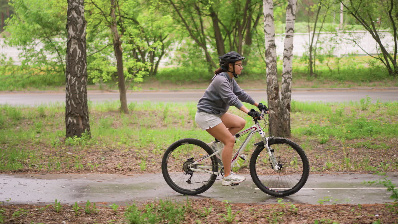 Woman Pedaling Along Paved Park Path Beside Trees And Roadside, Steady Commuting Pace With Helmet And Shorts, Leafy Foreground Framing, Healthy Outdoor Routine And Gentle Motion Captured Amid Green