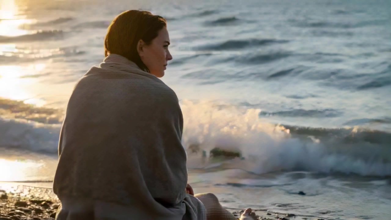 Sunlight reflecting water drawing woman wrapped blanket watching waves on pebble shore, copy space