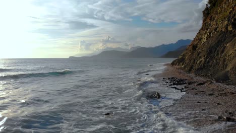 Sea-waves-splashing-on-rocky-coastline-after-storm-with-mountains-and-clouds-background-in-Mediterranean