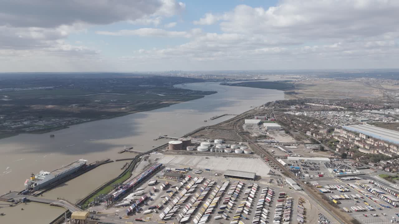 High-altitude sweep over Thames logistics corridor of cranes, containers and fuel tanks in Thurrock fading toward distant London skyline, depicting regional freight infrastructure and urban linkage