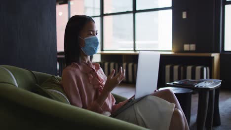 asian woman wearing face mask having a video chat on laptop at modern office