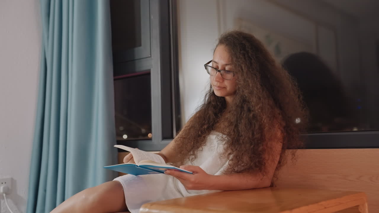 Femme écrivant dans un carnet d'hôtel près de la fenêtre, lumière douce d'une lampe, planification et journal en fin de soirée, posture pensive, stylo sur la page, notes de voyage et inspiration tranquille dans une chambre compacte