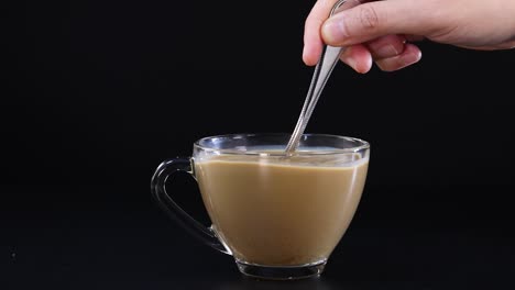 a hand stirs a coffee and milk mixture in a glass cup against a black background