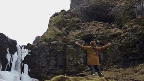 Tourist-hold-wide-open-arm-and-enjoy-Icelandic-landscape-with-waterfall-and-bird