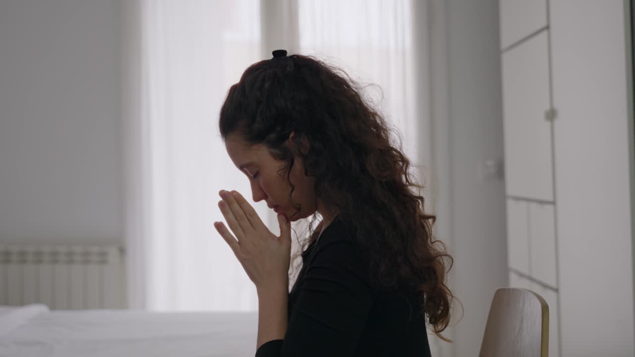 Religious young woman kneeling in bedroom and praying. Light shining through window curtain in background. Serious with hands clasped and eyes closed she is thinking and wishing inside room