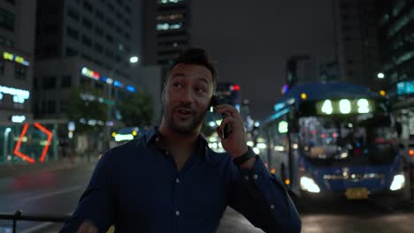 Cheerful-successful-young-businessman-talking-on-phone-about-his-great-sales-while-waiting-for-transport-at-bus-stop-in-Gangnam-street-of-Seoul