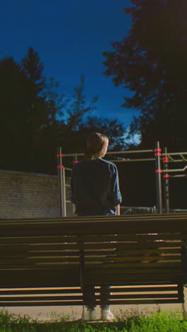 vista trasera de una mujer sentada al aire libre en un banco por la noche con la luz reflejándose suavemente en su cara, el fondo incluye una estructura de patio de recreo y una pared de ladrillo sombreada bajo un cielo azul profundo