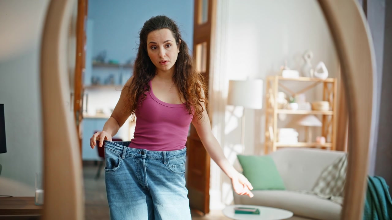 Fit woman examining oversized denim jeans in mirror home. Confused slim brunette
