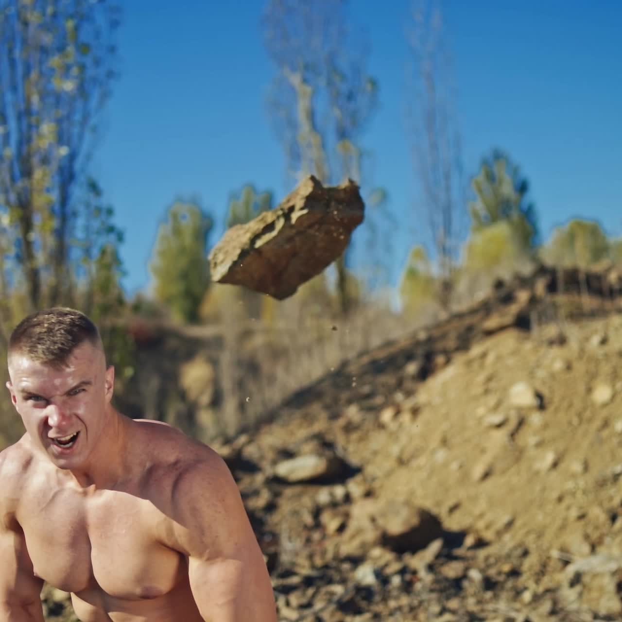 Athletic man exercises with stones. Strong sportsman taking heavy stone and throwing it over the head behind the back on rocky background. Slow motion.
