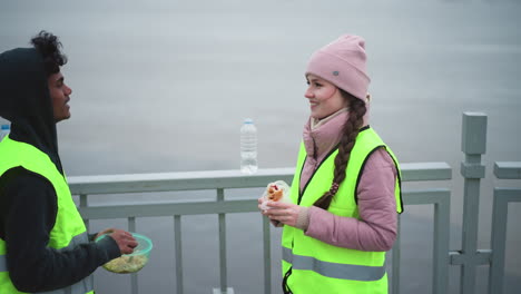 Volunteers in high visibility vests taking break on bridge during cold weather, standing by railing, holding food and water, talking and resting after outdoor cleanup near river with ice