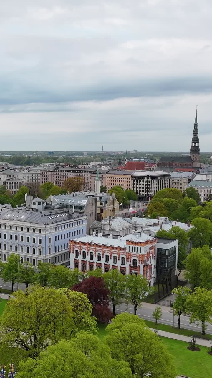 Riga Art Academy’s ornate façade with red and white Neo-Renaissance details stands by Esplanade Park, with St. Peter’s Church spire and city blocks in the background, vertical drone orbiting