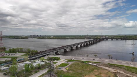 aerial drone shot of rem train crossing a bridge leaving the island of montreal traveling over the st lawrence river. blue overcast sky and urban landscape.