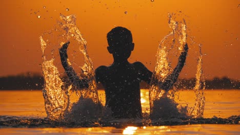 silhouette of boy at sunset in river creating splashes of water with his hands. slow motion