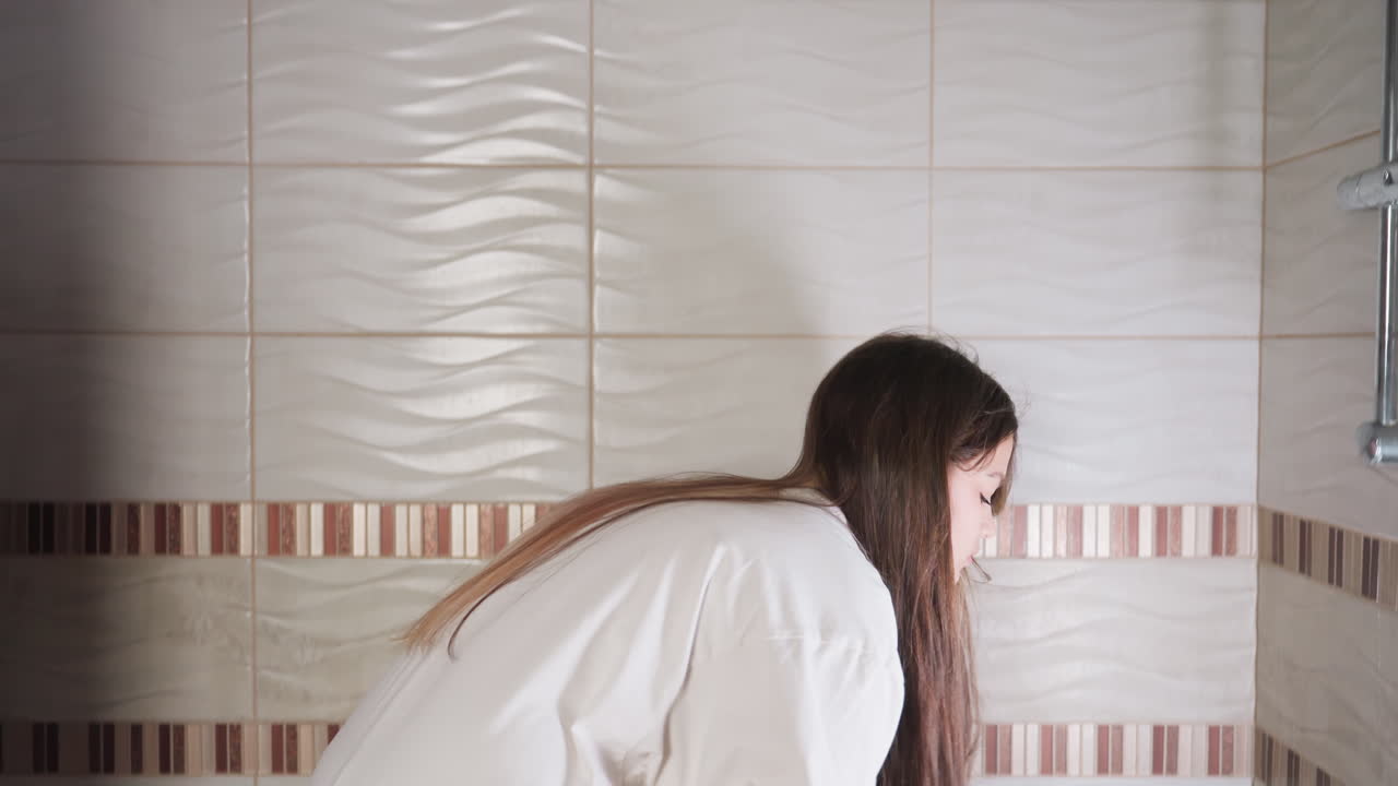 Young girl enters bathroom, walks to bathtub, pulls handheld shower, sits on tub edge near curtain, soft daylight on tiles, routine hygiene moment, calm mood, quiet movement with gentle reflections