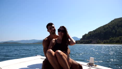 young pair sitting on bow of boat and posing to photographer at sunny day. happy couple in love spending time together on deck of ship and enjoying summer travel. concept of vacation or holiday. close up front view