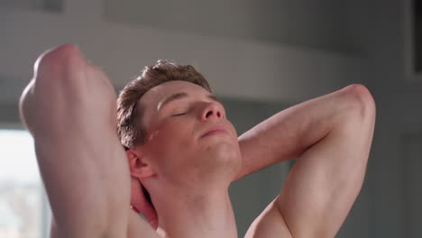 shirtless confident man with closed eyes stands in calm pose with both hands behind head, showing muscular arms and chest tattoo while expressing self-assurance in softly lit indoor room