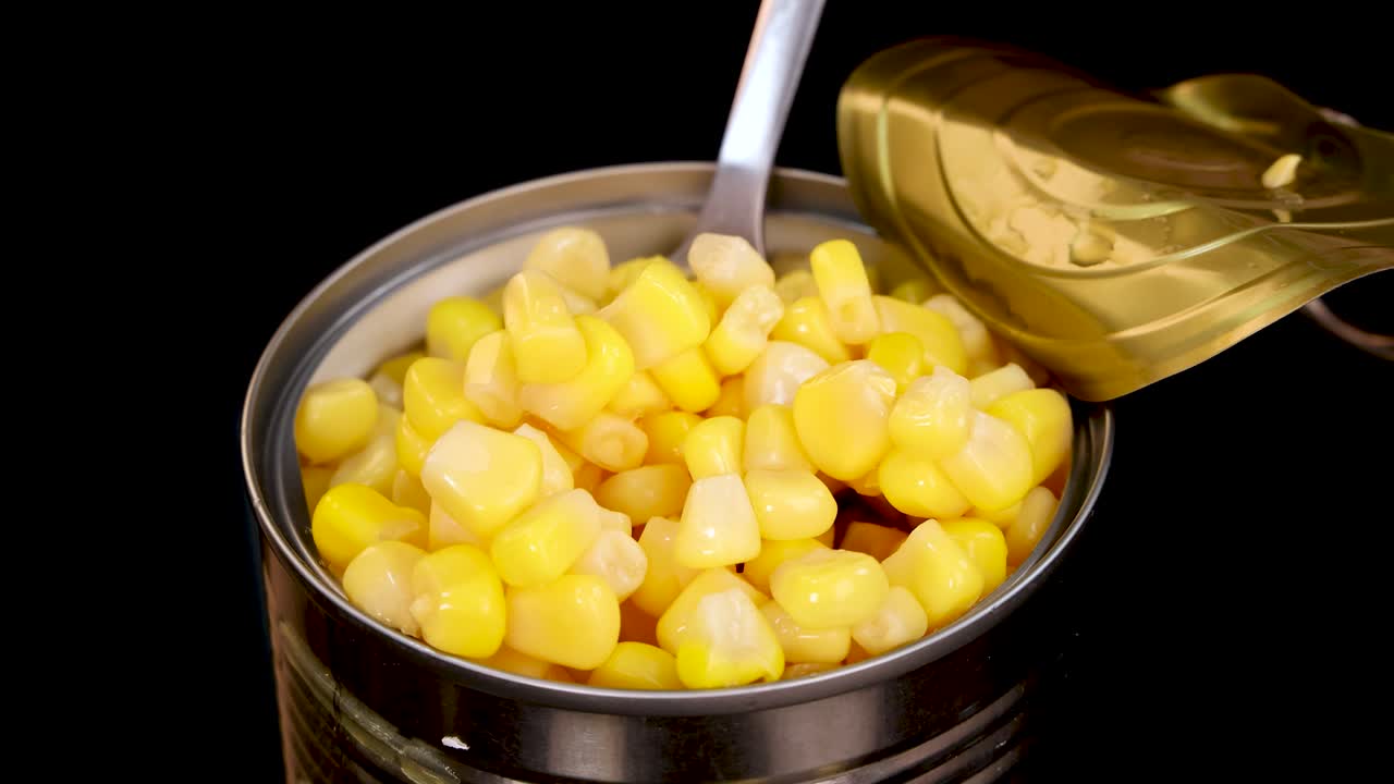 A metal spoon lifts yellow sweet corn kernels from an open tin can against a black background, with bright, even lighting and minimal camera movement