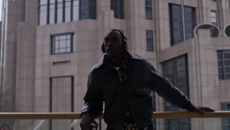 Black-Afro-American-freelancer-artist-photographer-looking-around-the-urban-metropolitan-cityscape-while-listening-to-music,-wearing-stylish-headphones