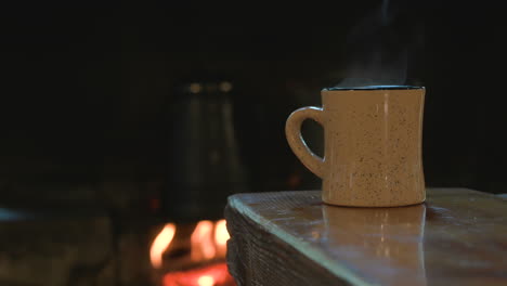 Steam-from-hot-coffee-rises-out-of-mug,-vintage-coffee-pot-over-fire-in-background