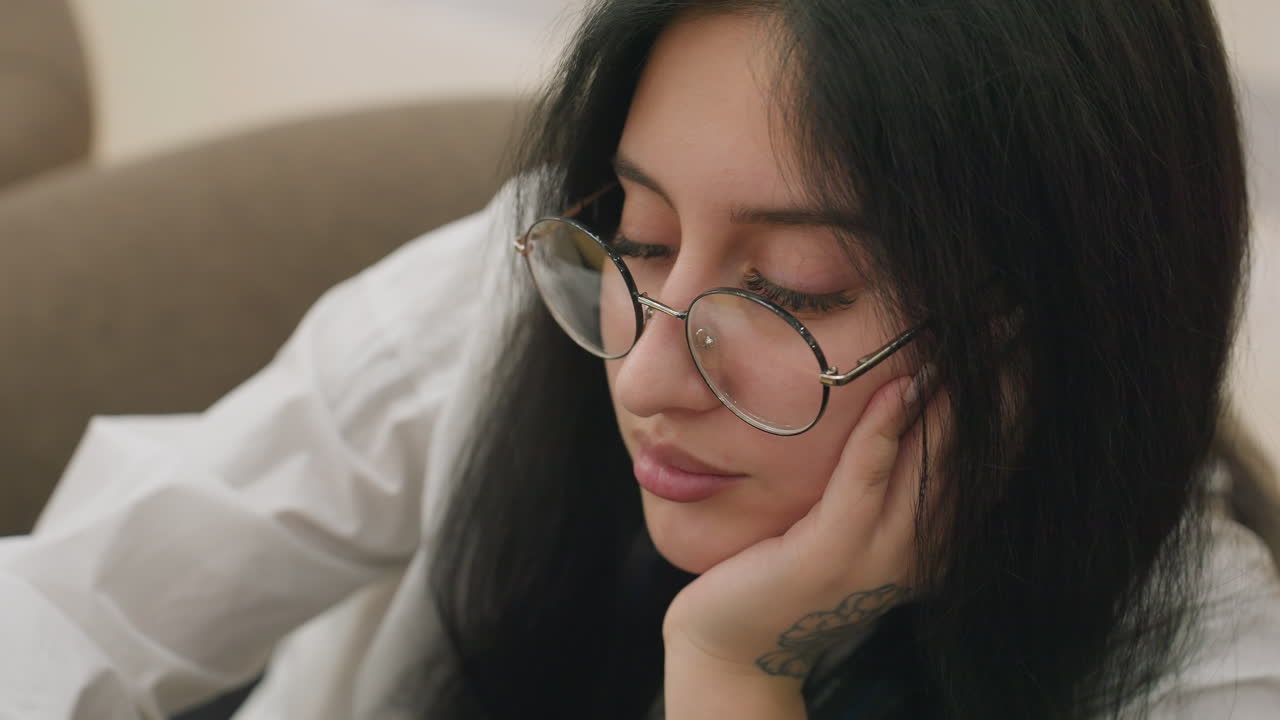Close up of professional lady wearing glasses resting head gently on hand with calm expression, seated indoors in soft lighting with visible hand tattoo and relaxed posture