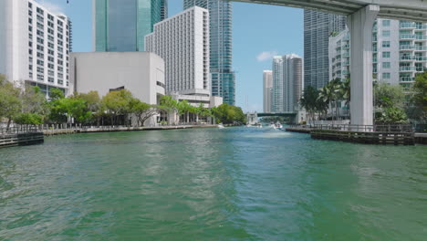 Forwards-tracking-of-motorboat-group-passing-under-bridge-in-modern-city.-Miami-river-and-downtown-buildings-on-waterfronts.-Miami,-USA