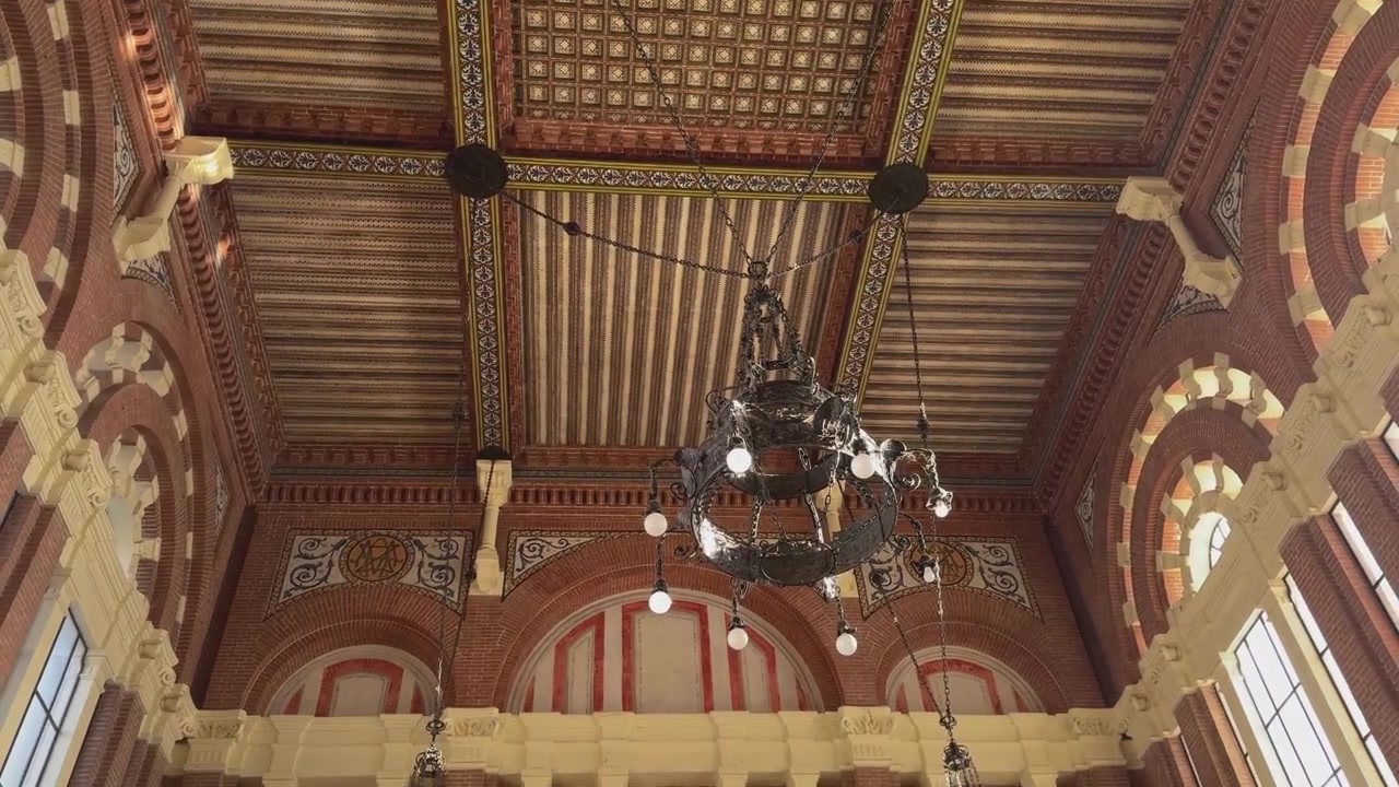 Handheld descending camera movement from the richly decorated Neo-Mudejar ceiling to the side walls of Aranjuez railway station, revealing arches, brickwork, white columns and architectural details.