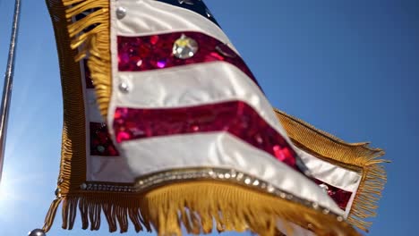 lujosa bandera estadounidense decorada con guiones y pañuelos brillantes, con franjas de color dorado, ondeando en el viento contra un cielo azul claro