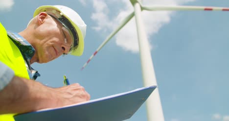 Engineer-Writing-On-Clipboard-While-Standing-Against-Windmill