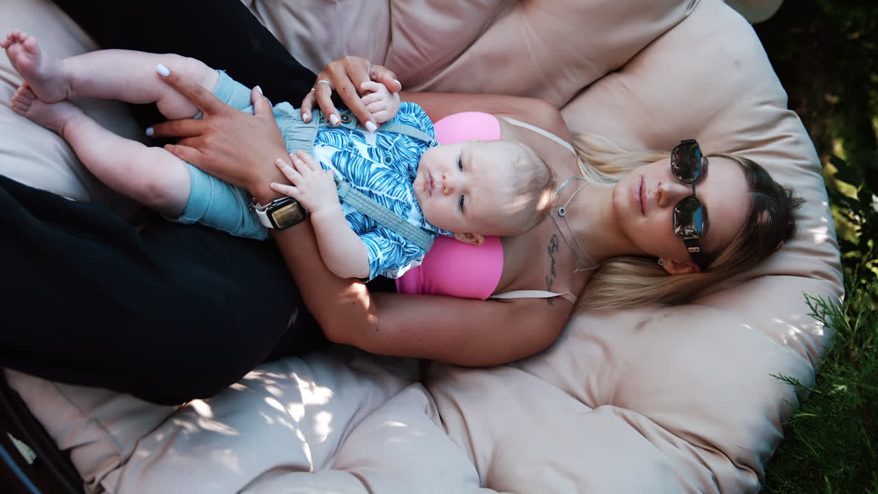 Beautiful blonde smiling lady laid back in a comfortable armchair. Mother holding her infant son sitting outdoors. Vertical screen.