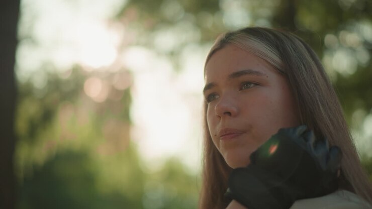 Beautiful young woman resting chin on her hand, gazing thoughtfully into the distance, sunlight creates a warm halo effect around her while blurred greenery