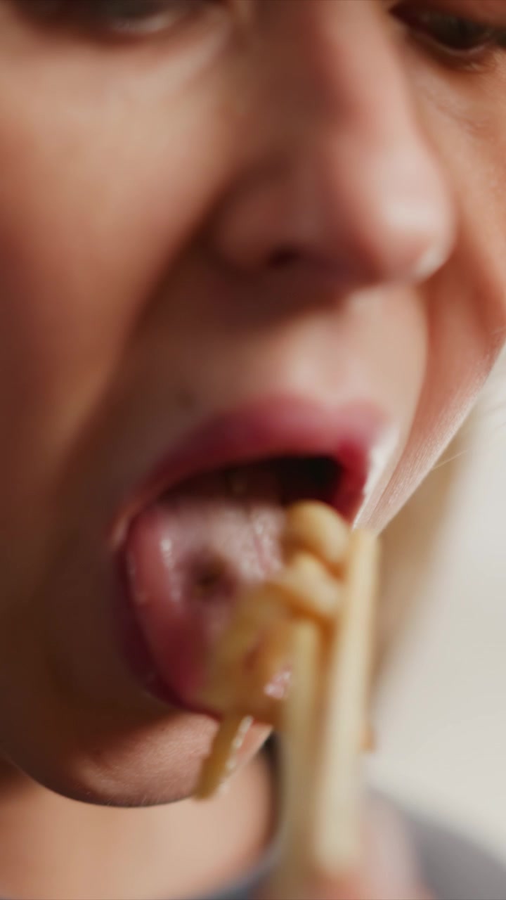 Potret dekat seorang wanita makan mi dengan sumpit, berfokus pada mulut dan ekspresi wajahnya. Adegan ini menangkap detail tekstur pada pori-pori kulit saat wanita tersebut mendekatkan makanan ke bibirnya, menunjukkan sedikit